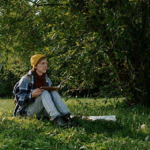 Person sitting on grass in a park, sketching in a notebook under a tree, wearing a yellow beanie and a plaid jacket, reflecting the outdoor adventure theme of Mystery Day Trips in Ontario.