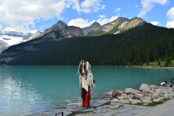 Person in traditional attire standing by a lake with mountains in the background