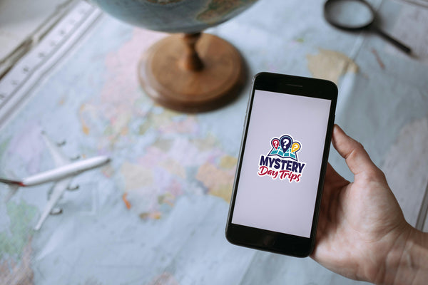 Person holding smartphone displaying the Mystery Day Trips logo, with a globe and map in the background, emphasizing curated adventure experiences in Ontario.